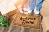 Man and Woman Unpacking Near Welcome Home Welcome Mat, Moving Boxes and Plant