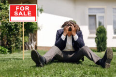 Depressed real estate agent sitting on grass in front of house for sale, outdoors